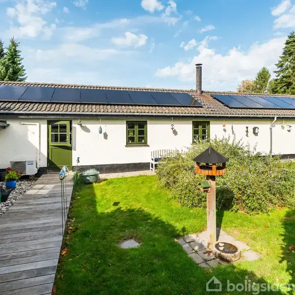 En hvid husbygning med solpaneler på taget står i en have med græs og buske. Der er en gangsti til venstre og et fuglehus i forgrunden.