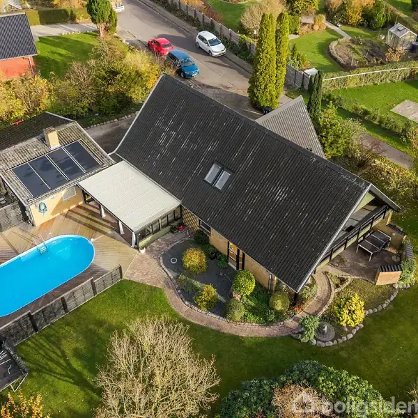 En moderne bolig med sort tag omgivet af en have. Ved siden af huset ses en terrasse med en oval swimmingpool indhegnet af et træhegn, i en rolig villavejsområde.