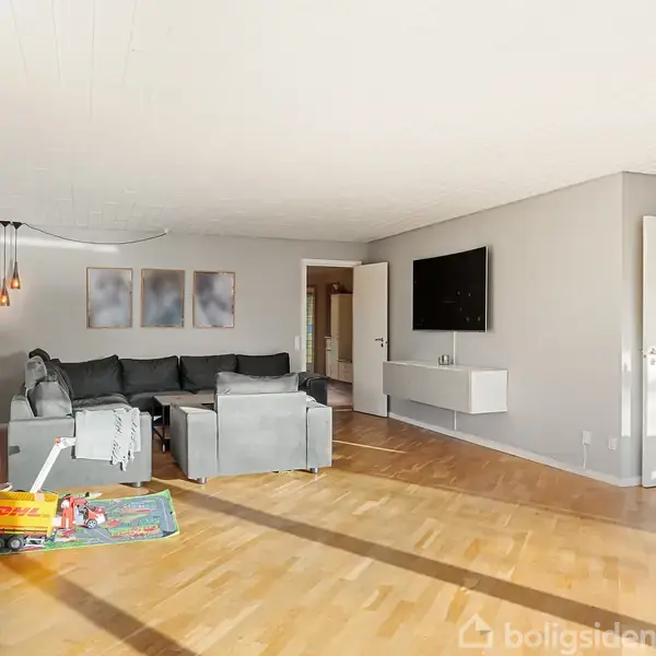 Grå sofaer står i en stue med trægulve og vægkunst. Et fjernsyn er monteret på væggen over en hvid hylde. Legetøj og bøger er opbevaret ved en reol.