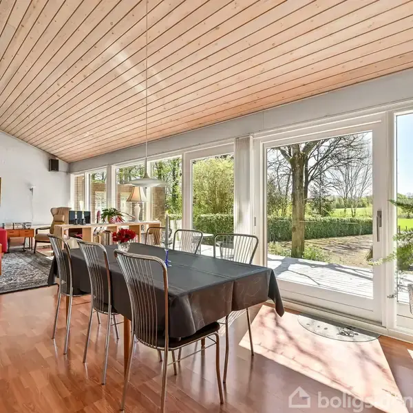 Spisebord med stole står i et lyst rum med store vinduer. Stuen inkluderer en rød sofa og udsigt til en grøn have. Træloft og gulv afslutter indretningen.