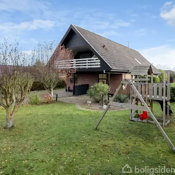 Hus med skråt tag står i grøn have; der er en legeplads med gyngestativ foran. Træer og buske omgiver området under en blå himmel med lette skyer.
