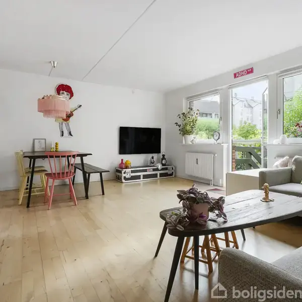 Stue med trægulv og sofa til højre, sofabord foran. Fjernsyn på væg i baggrunden. Spisebord med stole til venstre. Store vinduer giver udsigt til have.