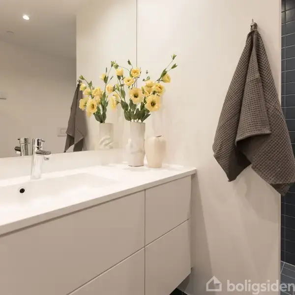 Hvidt badeværelsesskab med vask og spejl. En buket gule blomster i vase står på bordet. Hængende grå håndklæder ved siden af. Moderne indretning.