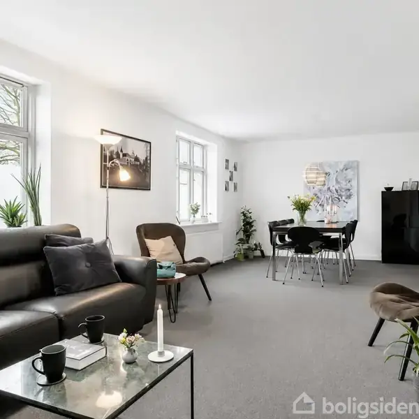 En moderne stue med en sort lædersofa og to stole, indrettet med et lille sofabord med to kaffekrus. Et spisebord med seks stole står i baggrunden. Lyse vægge med kunst og store vinduer.