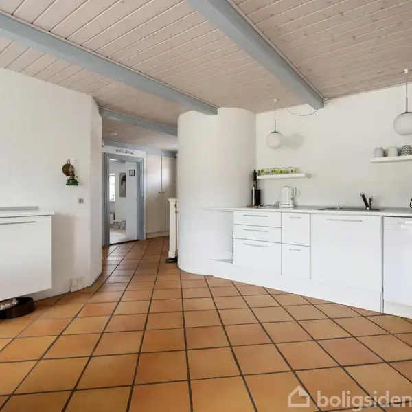 Køkken med hvide skabe og integrerede apparater, herunder opvaskemaskine. To hængelamper hænger over bordpladen. Væggene er hvide, gulvet er flisebelagt i terracotta. Maleri hænger på væggen.