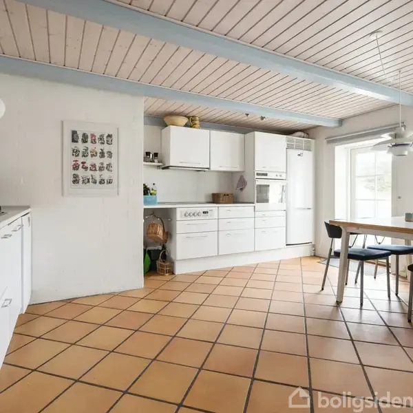 Køkken med hvide skabe står i et rum med lyse klinkegulve. Et spisebord med stole står ved et vindue. Hylde med pynt genstande er placeret på væggen.