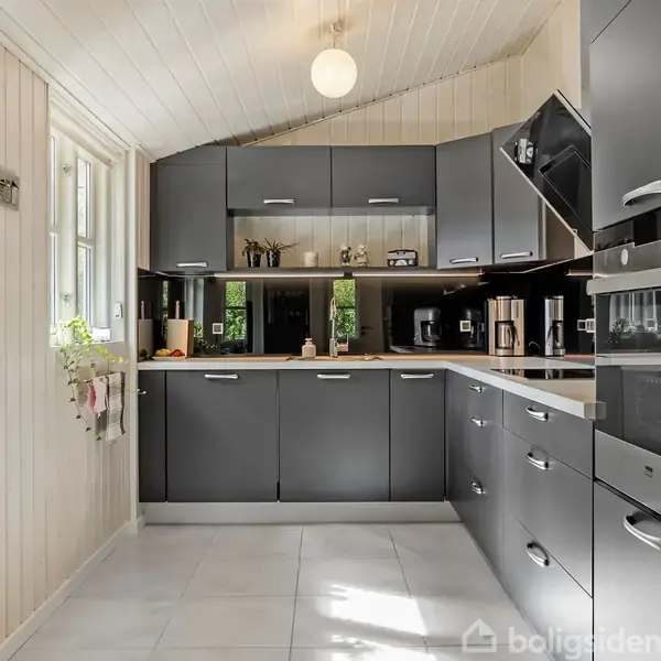 Et moderne køkken med sorte skabe i en hjørneopstilling, alt udstyret med indbyggede apparater. Detaljerne inkluderer vægpaneler, et stort vindue til venstre, og lys fylder rummet.