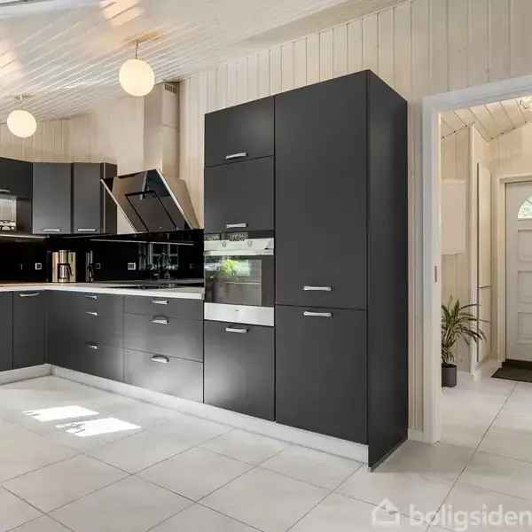 Moderne køkken med sorte skabe og hvide bordplader i et lyst rum. To runde lamper hænger fra loftet. En åben dør leder til en entré med en plante.