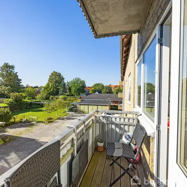 Balkon med klapstol og bord set udefra, rækker ud over grøn have i forstadsområde. Klare blå himmel og sollys oplyser området omkring villaer og små bygninger.