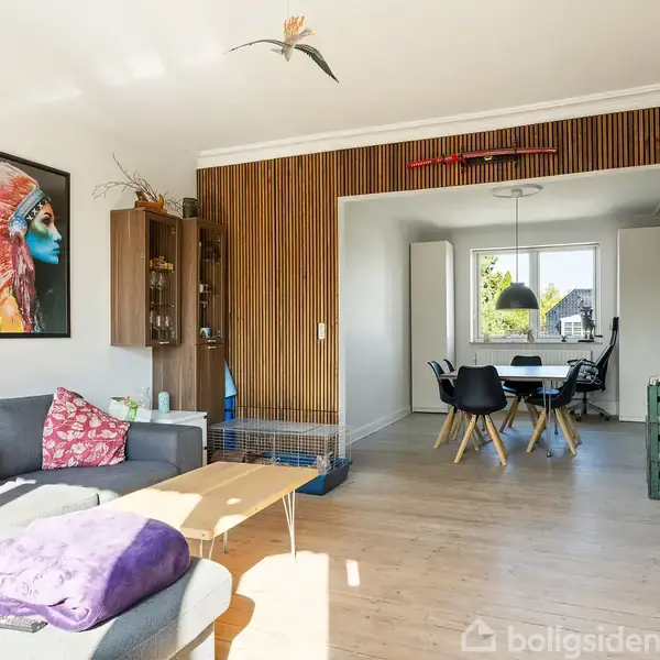 En stue med grå sofa har farverigt maleri på væggen og sofabord i træ. Rummet er lyst, med spisebord og stole i baggrunden. Der er puder i sofaen med teksten “Home is your happy place”.