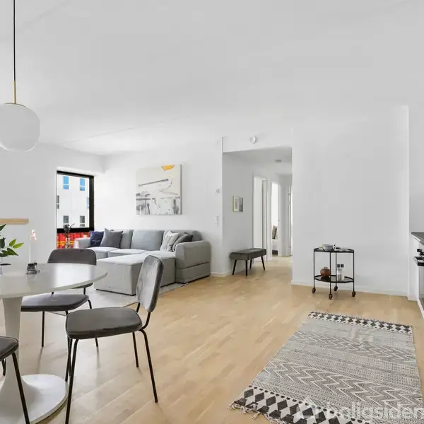En moderne stue med et rundt spisebord og fire stole står i forgrunden, mens en grå sofa med puder er placeret bagerst. Rummet er lyst med hvide vægge og trægulv.