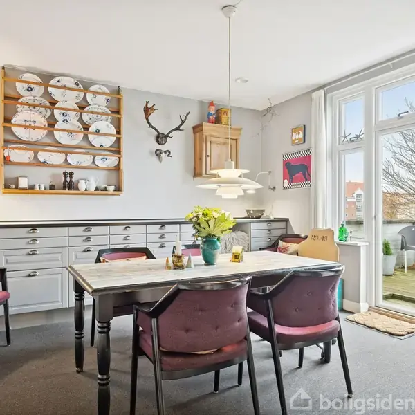Et spisebord omgivet af lilla polstrede stole står centralt i et lyst rum. Vaser med blomster pryder bordet, mens tallerkener er udstillet på en væg. Store vinduer åbner op til en altan.