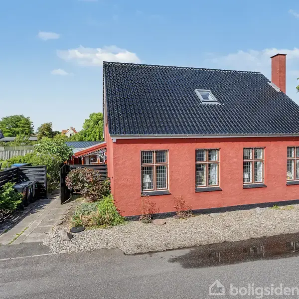Rødt hus med sort tag står ved en vej omgivet af grønne planter og en indkørsel. Klar himmel i baggrunden.