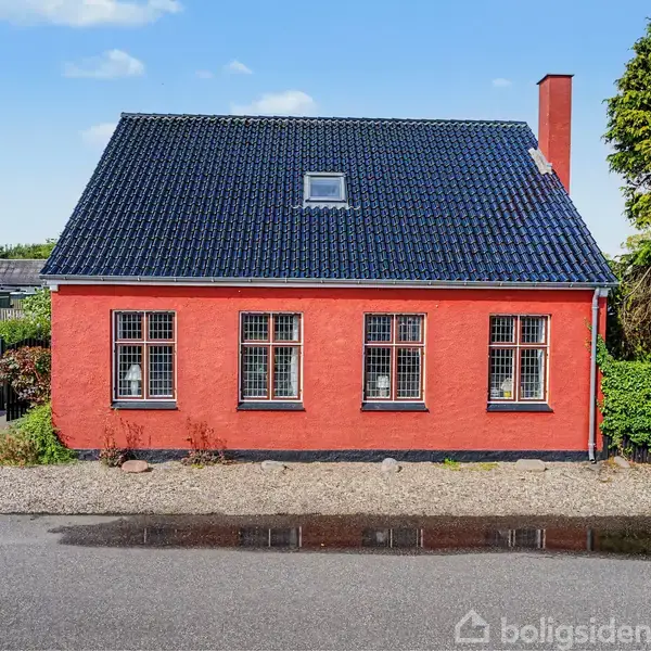 Rødt hus med sort tag står stille langs en vej, omgivet af buske og træer i et boligområde. Der er flere store vinduer foran.