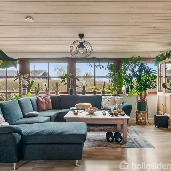 En stor L-formet sofa står foran et sofabord med pynt. Store vinduer bagved lader lys ind, og grønne planter dekorerer rummet. Trægulv og loft skaber en hyggelig stemning.