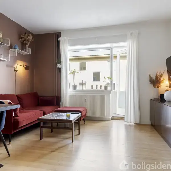 Stue med rød sofa står ved et sofabord; vægmonterede hylder indeholder pynt. Spisebord og stole til venstre, bogreol til højre. Store vinduer giver naturligt lys.