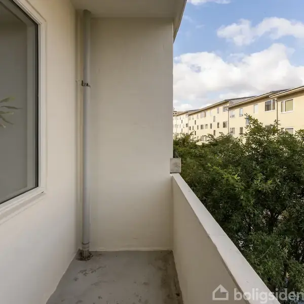 Balkon med hvidt rækværk og vægge. En plante ses gennem et vindue. Udsigt til grønne trætoppe og lysegule bygninger under en blå himmel med skyer.