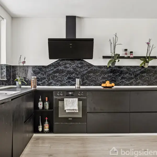 Moderne køkken med sort køkkenbord og vægmontering. Sort emhætte hænger over komfuret. Frugtskål og orkidéplanter pynter. Hylder med krydderier og glasflasker. Store vinduer til venstre.