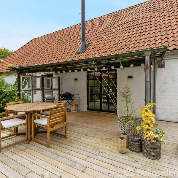 Et trædæk med runde træmøbler står foran et hvidt hus med rødt tegltag. Huset har sorte rammevinduer og en skorsten. Omgivet af en grøn have.