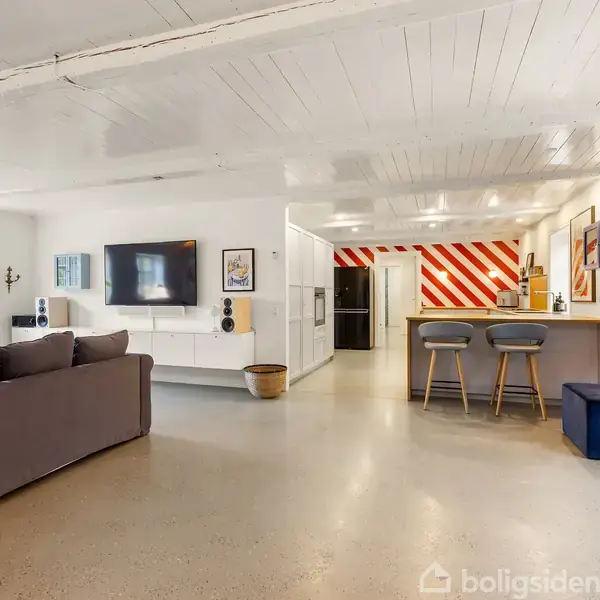 En moderne stue med grå sofa står mod venstre. Et TV hænger på en hvid væg. Til højre er en køkkenø med barstole og rødstribede vægge i baggrunden.