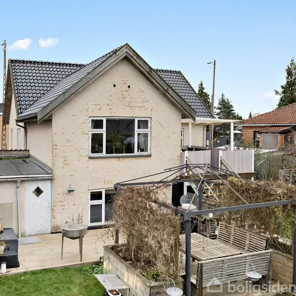 Et to-etagers hus med skråtag er omgivet af en have. Der er en terrasse og havemøbler under en pergola. Naboer og buske ses i baggrunden.