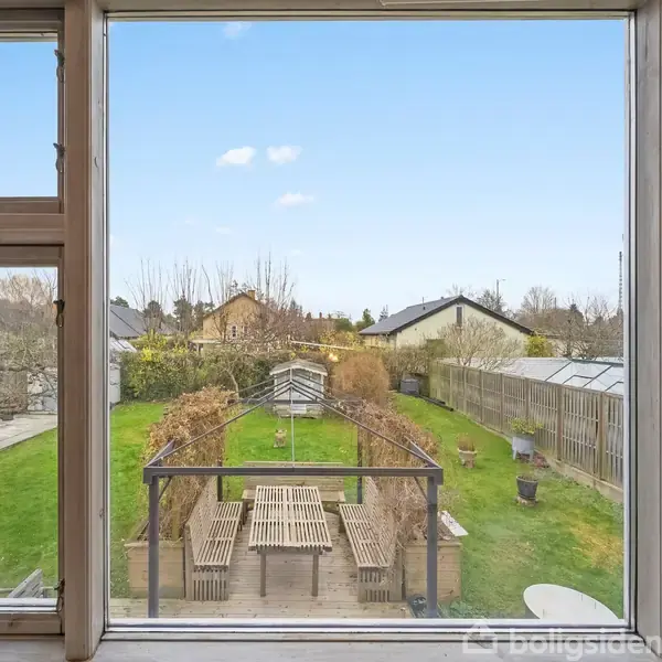 Et stort vindue viser en have med et langt træbord og bænke under en pergola. Omgivelserne består af græsplæne, hegn og træer i baggrunden.