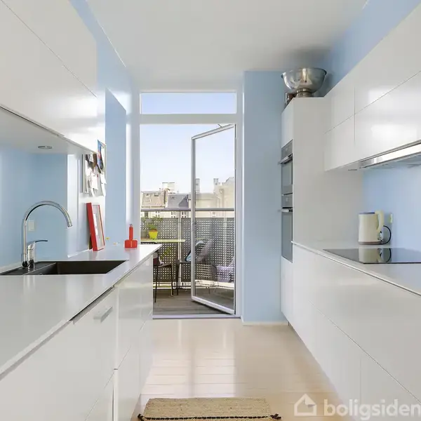 Lys køkken med hvide skabe og en sort vask, åben glasdør fører til altan med udsigt over bygninger. En elkande står på køkkenbordet.