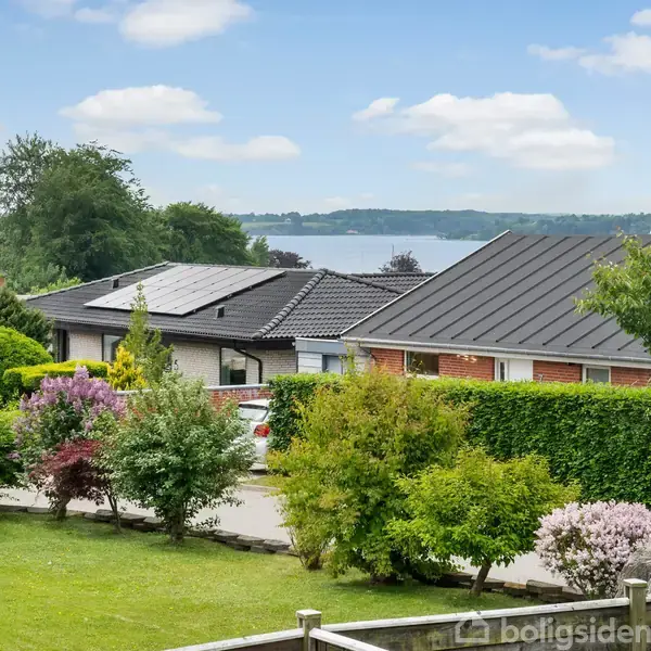 Et rækkehus med fladt tag omgivet af velplejede haver; i baggrunden ses en sø og træer under en blå himmel med skyer.