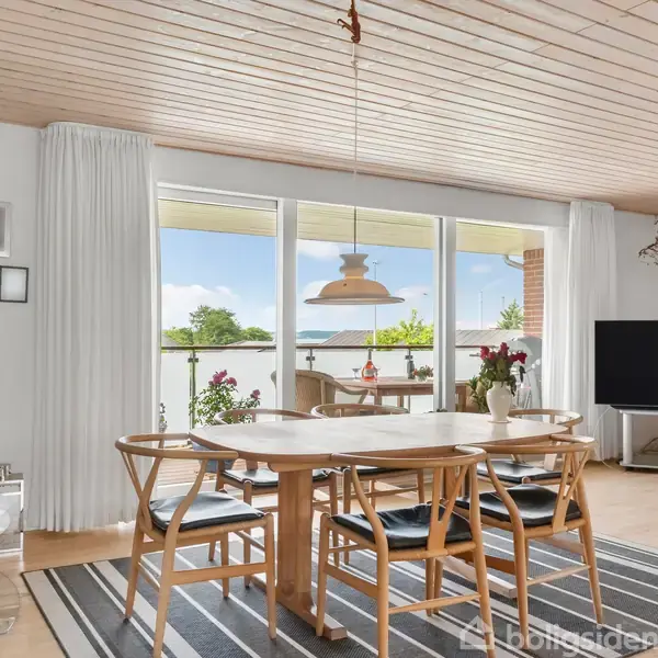 Køkkenø med grå bordplade, kogeplade og emhætte. Knive hænger på væggen. I baggrunden spisestue med store vinduer, udsigt til træer og himmel.