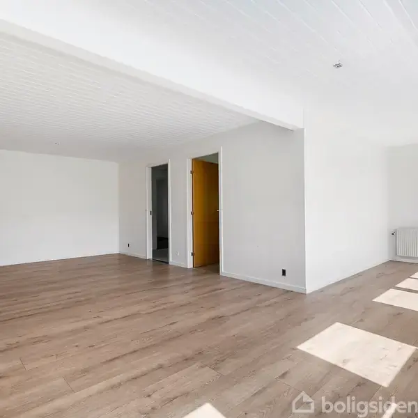 Et lyst, tomt rum med trægulve og hvide vægge åbner mod en lysfyldt krog ved et stort vindue. En radiator er placeret i hjørnet, og der er flere døre i baggrunden.