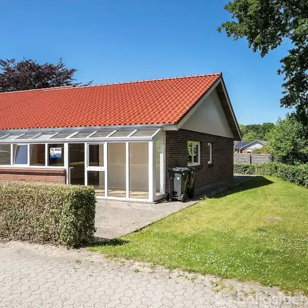 Et rækkehus med rødt tegltag og glasoverdækket terrasse, står ved siden af en trimmet græsplæne og flisebelagt indkørsel. Landsbymiljøet er grønt og fredeligt. Skilt i vinduet: "Til Salg".