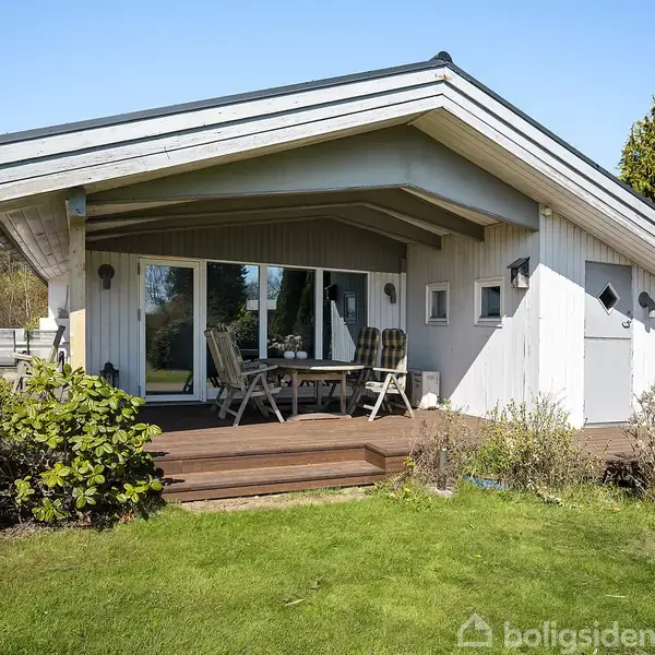 Et lille, lyst træhus med terrasse viser et bord og stole på verandaen. Huset er omgivet af grønne buske og græsplæne under en klar blå himmel.