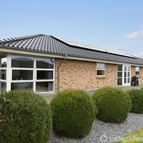 Et muret hus med tagsten står stille omgivet af en velplejet have med buske og grussti. Solpaneler er monteret på taget under en blå himmel med få skyer.