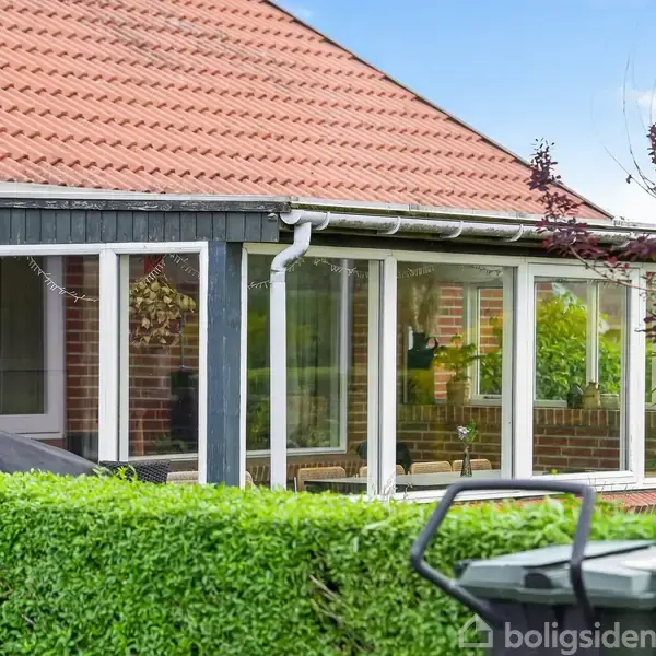 Et hus med rødt tegltag og en glasinddækket veranda. Omkring verandaen ses en grøn hæk, og et par skraldespande står i forgrunden.
