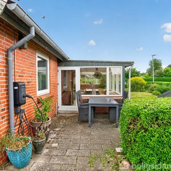 Rødt murstenshus med nummer 21 vises ved en lille gårdhave med fliser og bord. Krukker med planter står ved siden af huset. Baggrund med grøn have og buske.