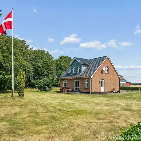 Et rødt murstenshus med sort tag står på en stor græsplæne. Et flag med rød og hvid farve vajer fra en flagstang. Omkranset af træer og marker.