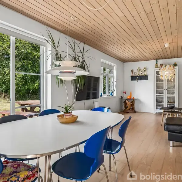 Et spisebord står i et lyst rum med træloft, omgivet af blå stole. Store vinduer til venstre åbner op til en have. Moderne indretning præger atmosfæren.