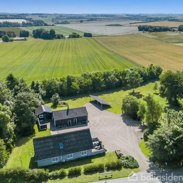 Et landsted med to bygninger ligger omgivet af frodige marker og træer. Bygningerne har mørke tage og hvid facade. Omkring ejendommen er der åbne, grønne områder under en klar blå himmel.