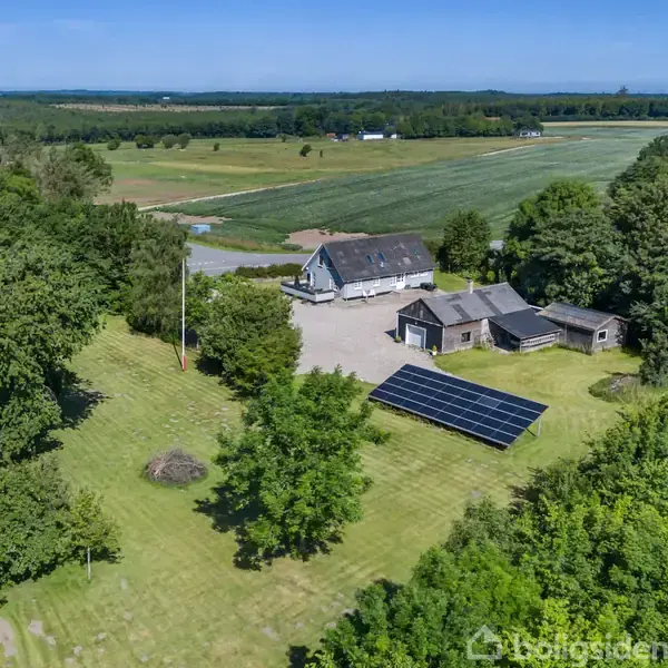 Et bondehus ligger i et grønt landskab med åbne marker omkring, omkranset af træer. Solpaneler er placeret på græsset tæt på husets udhuse.