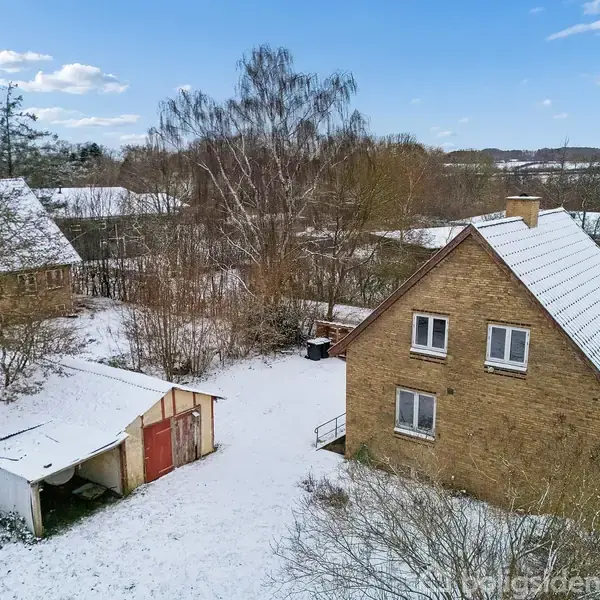 Et murstenshus med sneklædt tag står stille i et vinterlandskab, omgivet af træer og en snedækket grund. Et åbent skur ved siden af huset er delvist dækket af sne.