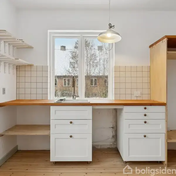 Et køkken med hvidt skab og træbordplade, tomt hyldesystem og vask under et vindue med udsigt til vintertræer. Flisebeklædt væg og trælamelgulv skaber en minimalistisk atmosfære.