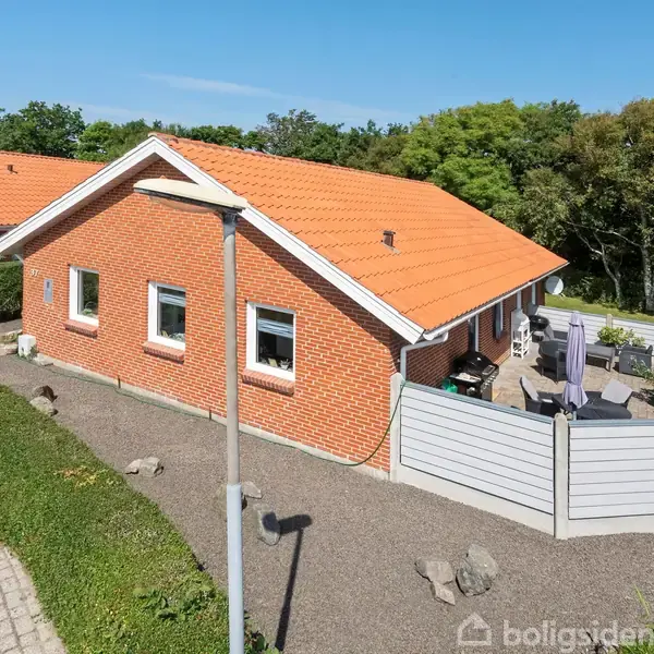 En rød murstensbungalow med orange tegltag står stille i et boligområde. Omkranset af grønne hække og træer, ses en indhegnet terrasse med havemøbler på højre side.