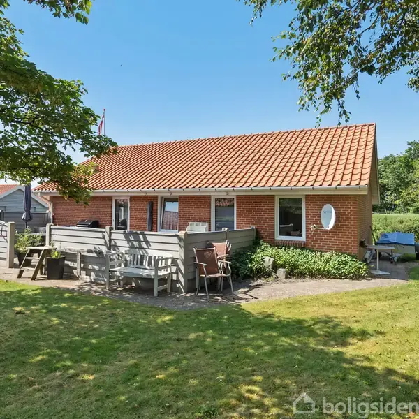 Rødt murstenshus med rødt tegltag står i en grøn have. Havemøbler er placeret på en terrasse foran huset. Baggrunden indeholder tæt bevoksning og træer.