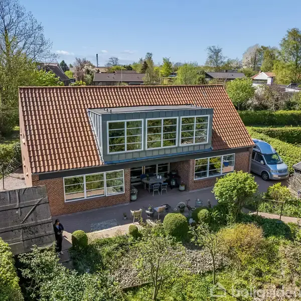 Et murstenshus med rødt tegltag har store vinduer med hvide rammer. Foran huset er der en terrasse med havemøbler. Haven er omgivet af grønne buske og træer i et boligområde.