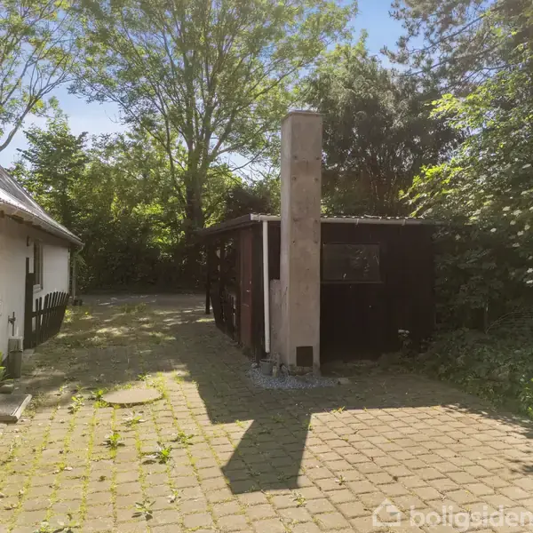 Et lille sort skur med en skorsten står i en gårdsplads ved siden af en hvid bygning. Omkring er der frodig bevoksning og træer. Ingen tekst er synlig.