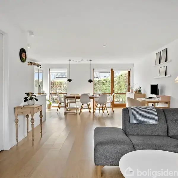 Grå sofa står i en lys stue, der har træmøbler, hængende lamper og en bordopstilling med stole. Store vinduer og glasdøre åbner ud mod en terrasse med grøn udsigt.