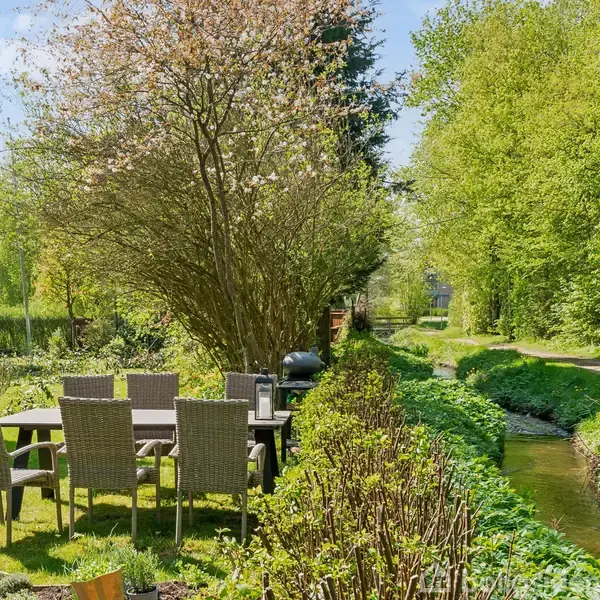 Et havesæt med seks stole står ved et bord på en græsplæne ved siden af et lille vandløb. Omkring er tætte, frodige grønne buske og træer under en klar himmel.