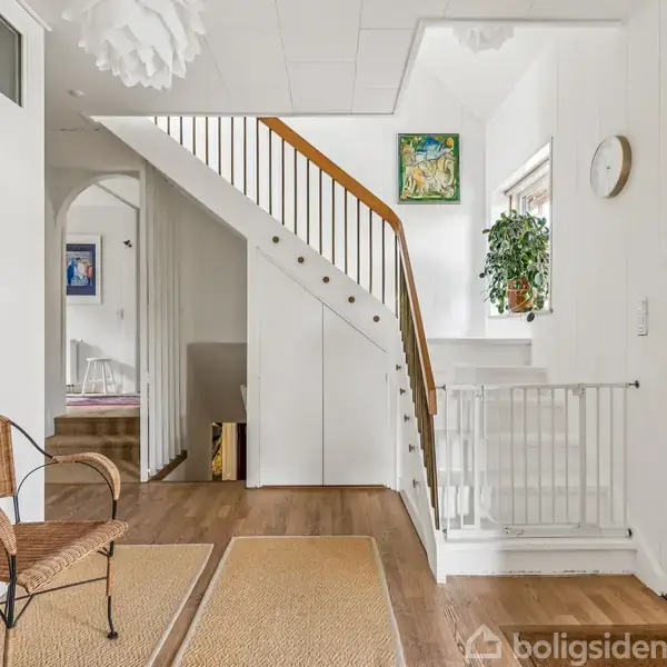 Trappe med hvidt gelænder fører opad i en lys entré. På væggen hænger farverige malerier. En plante står ved vinduet, og to tæpper ligger på trægulvet.