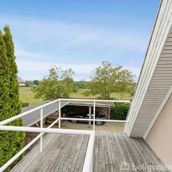En altan med trægulv og hvidt rækværk strækker sig fra et hus. Træer og en carport er synlige i baggrunden under en blå himmel.
