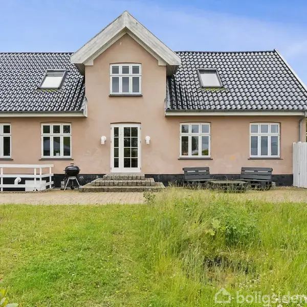 Hus med sort tegltag står i en velholdt have med græsplæne. Huset har store vinduer, en central trappeindgang og bænke ved indgangen. Sessile buske rundt om i haven.